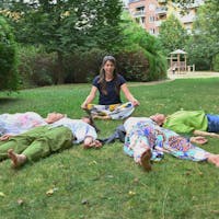Ariane gibt sitzend eine geführte Meditation im Park und die Gruppenteilnehmer liegen entspannt auf dem Gras
