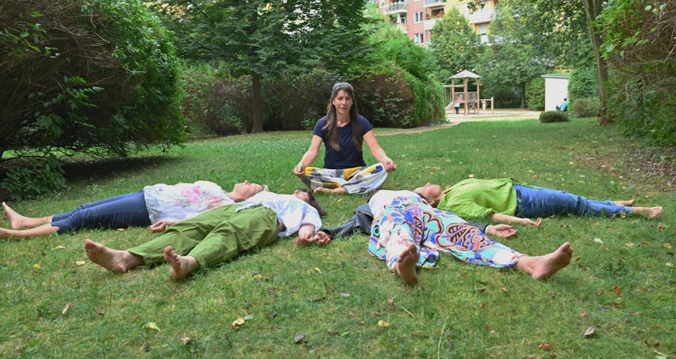 Ariane gibt sitzend eine geführte Meditation im Park und die Gruppenteilnehmer liegen entspannt auf dem Gras